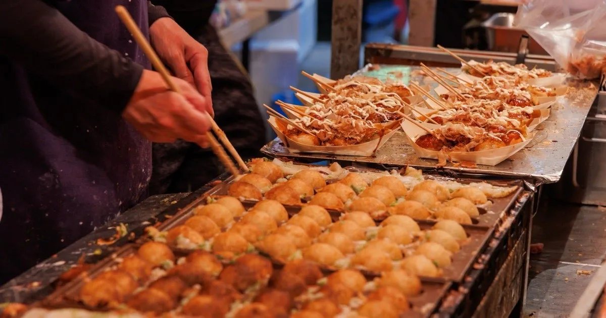Freshly made takoyaki on a hot griddle at an Osaka street stall, with finished trays topped with sauce, mayonnaise and bonito flakes
